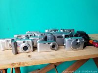 Photo showing multiple digital cameras arranged on a table, including models with silver and black bodies, various sizes, some labeled with megapixels (e.g., 10MP).