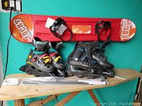 Photo shows two pairs of rollerblades, one red Lucky Bums snowboard, tent pole repair kit, and tent pole anchor spike arranged on a wooden table against a teal wall.