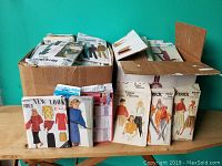 Two boxes filled with assorted sewing pattern packets from various brands on a wooden surface.