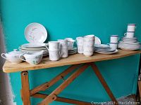 Photo of assorted dishware on wooden table showing white plates and mugs with gray floral designs, white plates with pastel floral patterns, and white dishware with blue stripe designs stacked on the right.