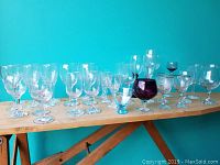 Photo showing the full assortment of 39 stemware pieces arranged in two rows on a wooden surface against a teal background, featuring clear glasses and a standout purple brandy snifter.