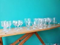 Wide view of all 30 assorted clear stemware pieces arranged in rows on wooden shelf showing different shapes and sizes.