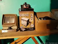 Photo showing vintage Super 8 film viewer, boxed slide and photo converter system, vintage camera on top, and box containing camera flashes.