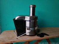 Front and right side view of stainless steel electric juicer with feed tube, locking arm, and pulp container. Black plastic accessory tool on table beside it.