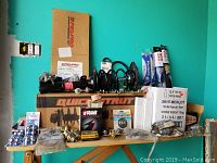 Overview of multiple boxed and loose NOS/NORS auto parts on wooden table against teal wall, showing various brands and types including strut assembly, wiper blades, carburetor gaskets, and fuel filters.