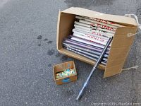 Cardboard box containing approximately 60 rolled posters, most wrapped in plastic, with one poster rolled outside the box. A smaller open box with poster mounting corners included.