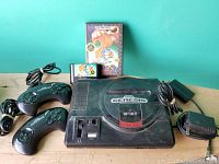 Front view of Sega Genesis console with two controllers, Sonic Hedgehog 2 cartridge and case, power adapter, and RF switch on table.