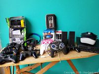 Full view of lot items arranged on a wooden table against a teal blue wall including gaming controllers, headphones, boxed accessories, PS4 and speakers.