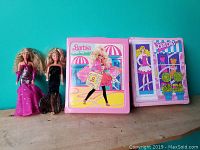 Two Barbie dolls placed standing in front of two pink Barbie carrying cases with graphic designs.