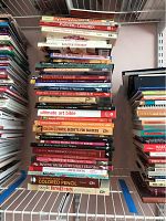 Overhead photo of a large stack of instructional art books on a white wire shelf, titles focusing on watercolor, acrylic, colored pencil, and mixed media techniques.