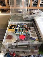 Photo showing stacked clear plastic storage containers filled with various scrapbook craft items including packaged stickers, paper shapes, and embellishments.