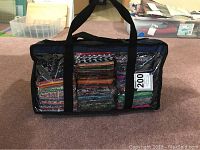 Closed clear vinyl carry case filled with bundled fat quarters, black fabric structure and handles visible