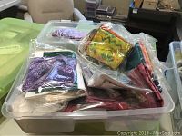 Clear plastic storage container filled with multiple pre-cut pieces of batik cotton fabric in clear plastic bags, showing various bright color patterns including purple, yellow, and orange.