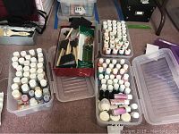 Wide shot of three clear plastic storage containers. Two containers filled with white-topped acrylic paint bottles in assorted colors. One container with smaller paint bottles and paint mediums along with a box of assorted paint brushes on top including foam and bristle brushes with wooden handles.