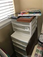 View of two white plastic storage bins stacked with an assortment of pastel sets on top including a wooden case and boxed pastel sets.