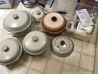 Seven cooking pots with lids, one extra lid, and a white plastic Salad Shooter with instruction manual visible in a plastic container.