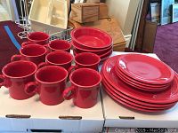 Photo showing the entire set of red Fiesta Ware dishes including mugs, dinner plates, small plates/saucers, and bowls arranged on white boxes.