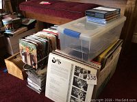 Wide view showing crates and boxes containing vinyl LPs and 45 RPM records, a clear plastic tote with blue latch, and DVDs stacked on top including Downton Abbey series.