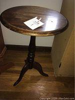 Front side view of the wooden accent side table showing round top with some wear and turned pedestal base standing on three feet.