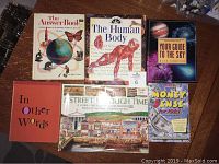 Overview of six children's reference books arranged on a wooden table, showing front covers including 'The Answer Book', 'The Human Body', 'Your Guide to the Sky', 'Money Sense for Kids', 'In Other Words', and 'A Street Through Time'.
