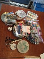 Photo showing multiple plastic bags containing threads, buttons, needle books, scissors, and various sewing supplies on a wooden surface.