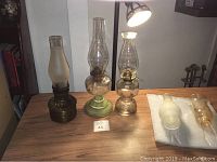 Three vintage oil lamps on wooden surface under lamp light, two glass and one brass metal with two extra hurricane glass tops on protective foam.