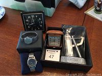 Photo showing six items on a table including two clocks (one in brown leather-like case and one black Westclox wind-up clock), two wristwatches on pillows, Timex watch band and attachment tool.
