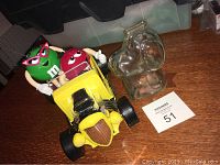 Photo showing the M&M's hot rod candy dispenser with clear glass Snoopy bank on a wooden surface, featuring two M&M character figures.