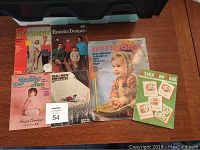 Six colorful vintage crochet and knitting booklet covers spread on table with green crochet hook package included, showing various patterns and designs for children and adults.