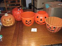 All five pumpkin items arranged on table: a light-up ceramic pumpkin decoration with plug, two pumpkin trick-or-treat buckets (one with a stuck battery compartment), one treat bowl, one pumpkin planter.