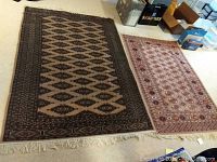 Wide angled photo showing two rugs side by side. Left rug is tan and black geometric design, right rug is tan with pink and beige floral motifs.