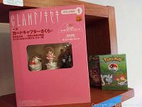 Boxed Clamp Exhibition of Works 1989-2004 Volume 1 with visible Cardcaptor Sakura figures and two Pokemon TCG boxes on wooden shelf.