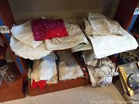 Multiple assorted linens stacked on shelves, including several placemats and folded tablecloths and napkins, mostly white or cream with embroidery, one red item visible