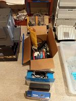 Box with multiple cables, surge protectors, and electronic items stacked showing a blue Linksys PSUS4 box and other items.