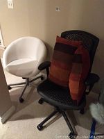 Black mesh Herman Miller Aeron chair and white swivel chair side by side with two striped pillows on black chair.