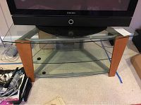 TV stand with a Samsung TV sitting on top, showing glass top and two glass shelves underneath. Wood legs and metal frame sides visible.