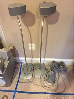 Two Bose satellite speakers mounted on tall silver stands with cables coiled at base and three more speakers with cables nearby on carpeted floor against wall.