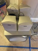 Full view of speaker system showing one subwoofer and four satellite speakers, all in off-white color with wires attached.