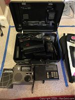 Photo of the open case with RCA VHSC camcorder, power adapter, and cables, Sony portable radio, Grundig shortwave radio and manual on floor surface.