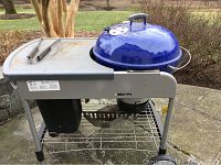 Side view of Weber grill showing blue dome lid, metal side table, tongs resting on side table, metal frame, and black bin below.