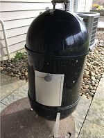 Full view of the Weber black vertical drum smoker with a white access door and metal legs, positioned outdoors on a mat with rocks and siding in the background.