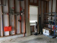 View of garage corner showing yard tools hung on wood wall mounts, ironing board, upright vacuum, and shelving unit with various items.