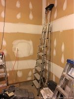 Full view of the aluminum step ladder standing against a drywall unfinished wall, showing step details and height.