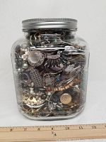 Full view of the glass jar filled with assorted jewelry pieces from one side with visible wide variety of metallic and beaded jewelry items.