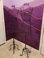 Full view of two black metal wire dress forms standing side-by-side against purple wall backdrop.