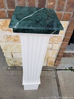 Front view of the full pedestal showing painted wooden base and green stone top against a brick wall background.