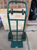 Front upright view of green metal dolly, showing two large black tires, handle with pin for adjustment, and top caster wheels.