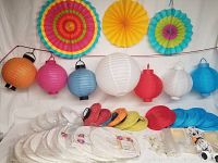 Photo showing three large colorful paper fan decorations arranged vertically, various paper and fabric lanterns in different colors including white, red, blue, pink, orange, lighter red, and light blue, two lantern cord kits and multiple lightbulbs, all arranged on a light background.
