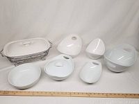 Seven white porcelain serving dishes arranged flat on a white surface with a ruler showing scale. Includes one oval dish with lid and metal stand along with varied shaped smaller dishes.