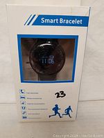Front of the box showing black smart bracelet with digital time display along with icons for call reminder, sleep monitoring, calorie consumption, movement distance, anti-lost alarm, and remote shoot.